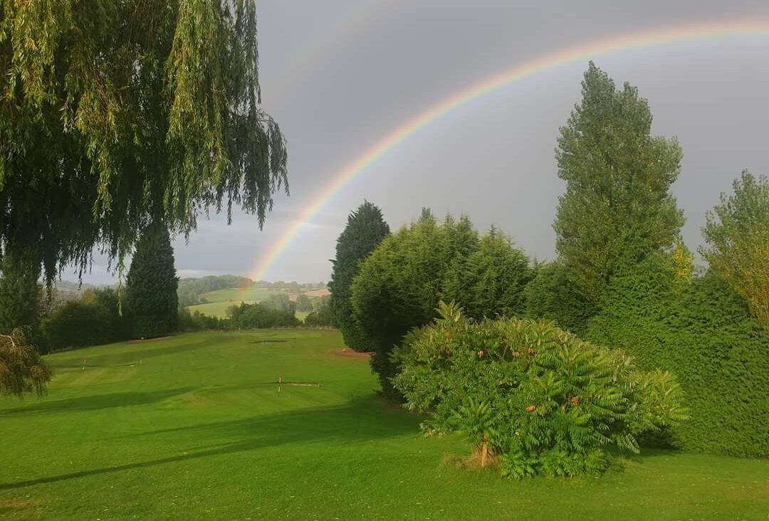 Green countryside surrounds the grassy view with a rainbow in the distance cutting through the sky.